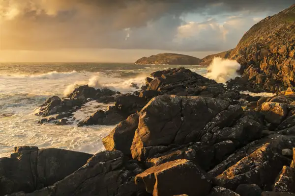 Rocky coastal landscape with waves crashing against dark, jagged rocks. The scene is bathed in warm, golden light from a setting or rising sun, casting long shadows. In the background, an ocean stretches to the horizon under a partly cloudy sky, with a distant landform visible.