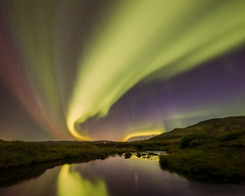 Nightscapes: Northern Lights in Iceland displaying a mesmerizing array of greens and purples that arc across a star-filled night sky, with their reflection shimmering on the surface of a tranquil river below. Gentle hills frame the scene. a sky view looking up at the camera