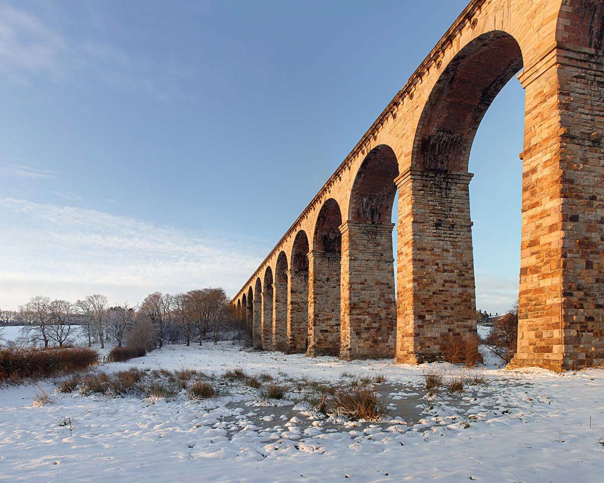 Sunlight illuminates the Crimple Viaduct in winter  a snow covered bridge in front of a brick building
