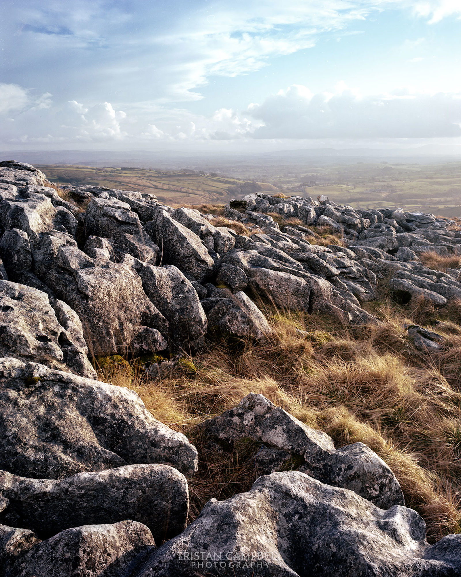 Rocky landscape with grey stone slabs and scattered yellow-brown grass under a partly cloudy sky. The background reveals a vast expanse of rolling fields under a gently lit, expansive horizon.