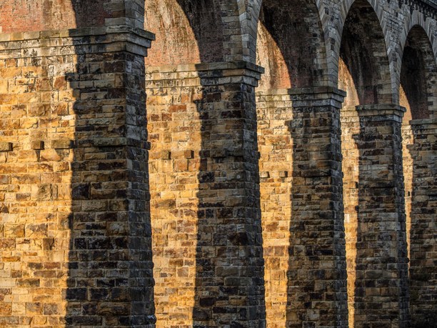 The Crimple Viaduct: The Crimple Valley Viaduct's stone structure bathed in sunlight. Arches rise from sturdy pillars, their shadows striping the textured walls. The golden hue of the stone contrasts with the soft arch shadows, evoking a sense of historic grandeur.