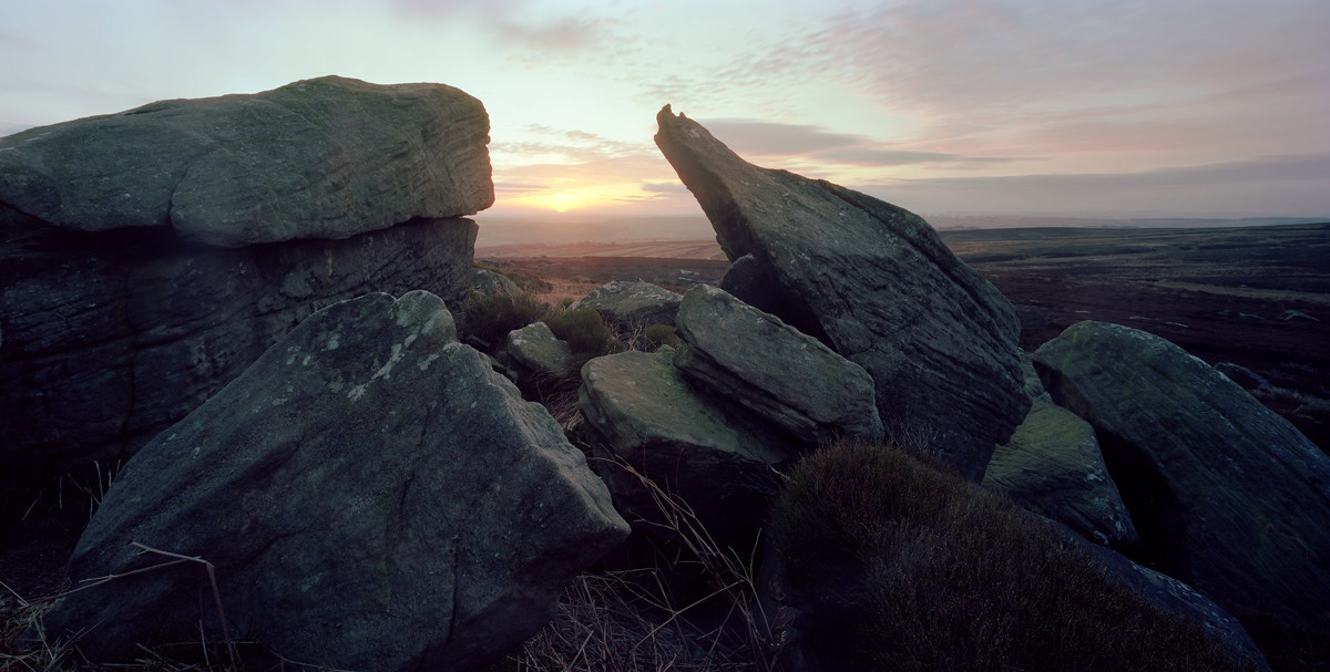 A twilight scene over North Yorkshire moors with rugged rocks in the foreground. A warm glowing horizon peeks through the stones, casting a gentle light over the expansive heather-clad landscape, which stretches into the distance. a view of a large rock