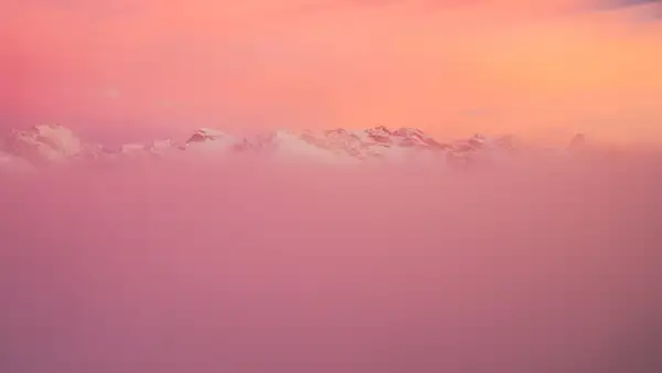 Snow-capped mountain peaks partially obscured by a layer of mist, with a vibrant pink and orange sky at sunset creating a serene, dreamy atmosphere.