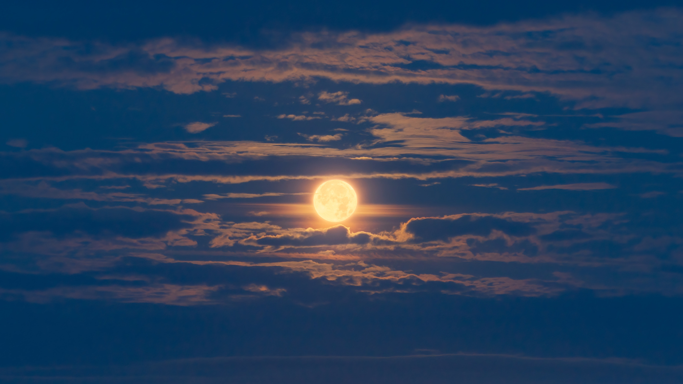 A full moon emits a glowing light through scattered clouds in a twilight sky. The moon's brilliance creates a halo effect, with the clouds illuminated in soft shades of blue and orange, evoking a peaceful nocturnal scene.