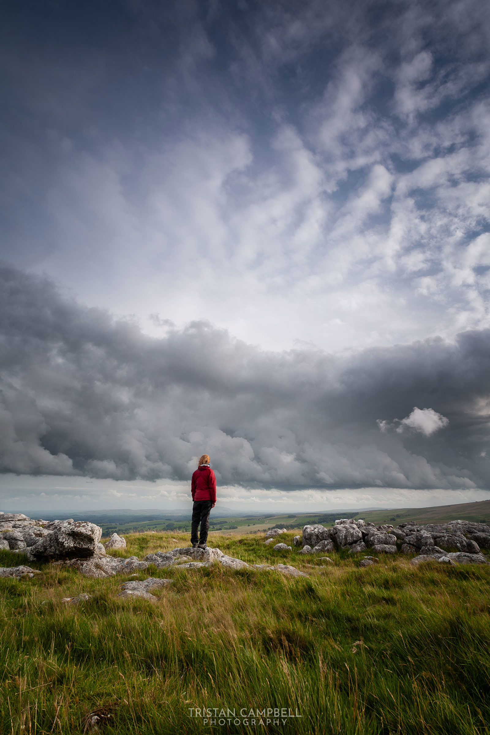 A person in a red jacket stands on a grassy hill with scattered rocks, looking out over a vast landscape under a dramatic, cloud-filled sky. The dark, rolling clouds dominate the upper portion, contrasting with the greenery below, suggesting an approaching storm.