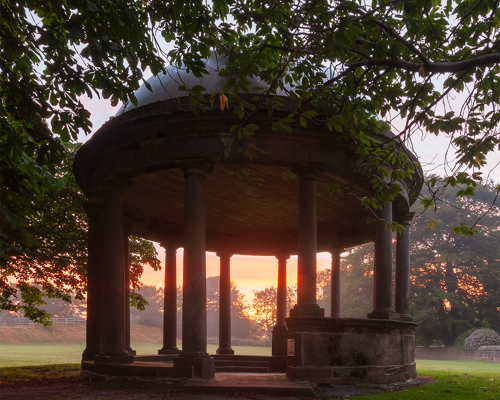 Harrogate Landscapes: A tranquil early morning at the Stray in Harrogate. A classical domed structure with stone pillars stands amidst the trees. Its silhouette is framed by the soft orange hues of sunrise peeking through the columns, contrasting with the structure's shadowy interior. Overhanging branches from nearby trees add a touch of greenery to the scene. a gazebo with columns and a tree in the background