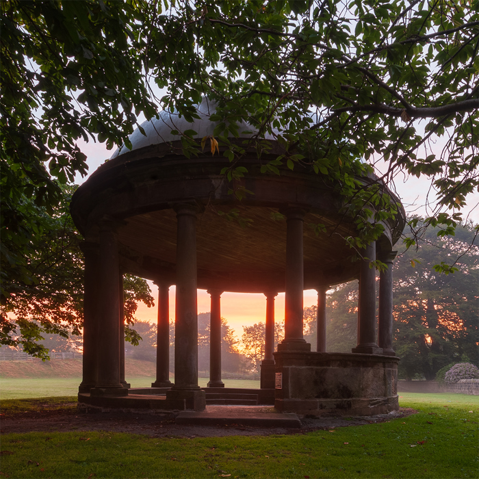 A tranquil early morning at the Stray in Harrogate. A classical domed structure with stone pillars stands amidst the trees. Its silhouette is framed by the soft orange hues of sunrise peeking through the columns, contrasting with the structure's shadowy interior. Overhanging branches from nearby trees add a touch of greenery to the scene. a gazebo with columns and a tree in the background