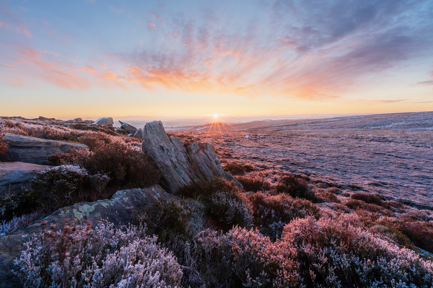 A vivid sunrise graces a frost-covered moorland with pink heather and rocks. A sunburst peeks above the horizon, casting a warm glow on the scene and painting the sky with hues of orange, pink, and blue. Frost glistens on the heath and rocks, suggesting a cold morning.