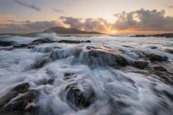 Waves cascade over dark, weathered rocks in the foreground, creating a misty, blurred effect. In the background, a softly illuminated horizon showcases a glowing orange sunset beneath a canopy of clouds. A distant silhouette of a hill rises on the left, partially obscured by the ocean spray.