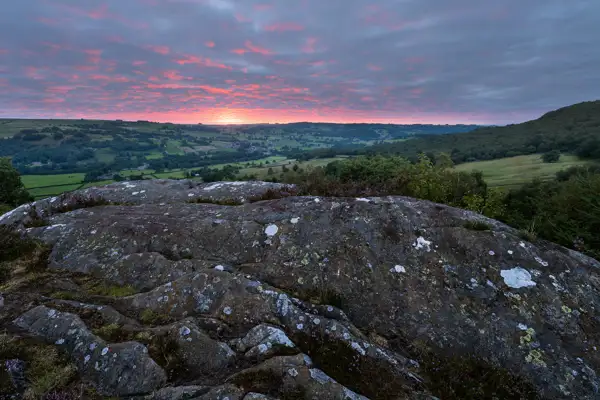 Rocky foreground with patches of lichen and small plants, overlooking a vast, lush green landscape of fields and rolling hills. A vibrant sunset casts pink and orange hues across a cloud-dappled sky, creating a dramatic horizon.