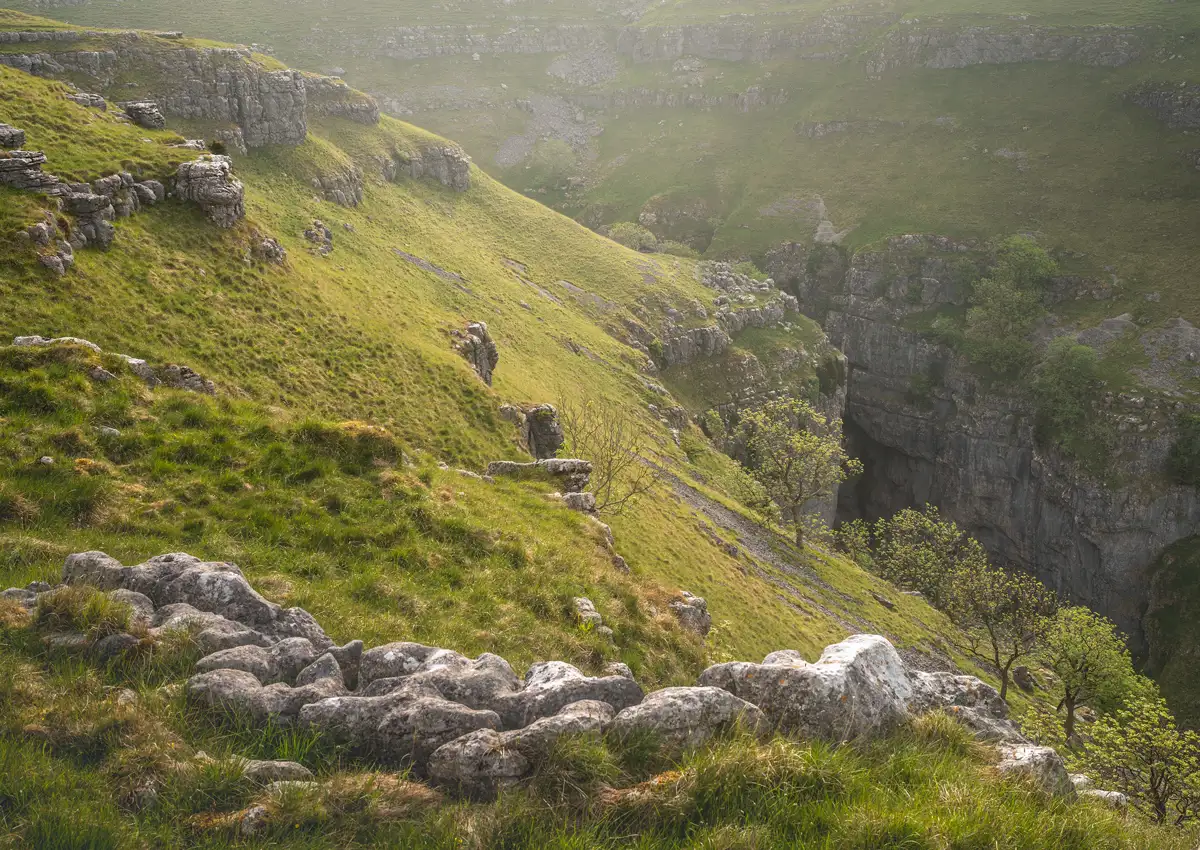 Sunlit rolling hills with patches of lush green grass and rocky outcrops. A steep rocky cliff drops into a shadowy gorge below, while scattered trees with fresh green leaves grow along the slope. The scene is bathed in soft, warm light, creating a serene and tranquil atmosphere.
