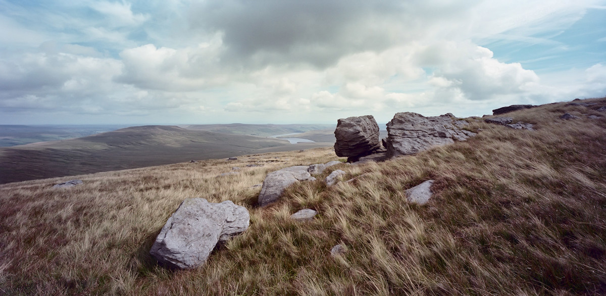 Rolling hills of North Yorkshire moorland under a vast sky with scattered clouds. Foreground shows tufted grass and scattered rocks, midground reveals undulating landscape, and a sliver of water nestles in a distant valley. a field with a mountain in the background