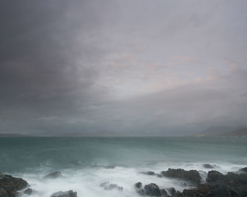 Seascapes of the Outer Hebrides: Moody grey clouds hover over churning turquoise waters. Mist-like waves crash against rugged rocks, blurring into a haze at the shore. The remote landscape exudes a serene yet powerful atmosphere. a group of clouds in the sky