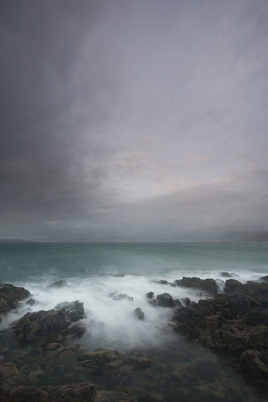 Moody grey clouds hover over churning turquoise waters. Mist-like waves crash against rugged rocks, blurring into a haze at the shore. The remote landscape exudes a serene yet powerful atmosphere. a group of clouds in the sky