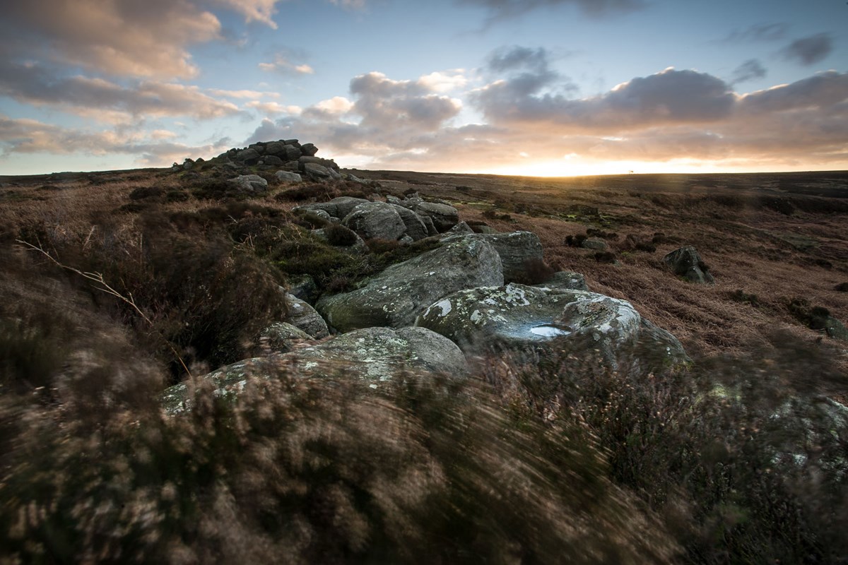 Outcrop on the North Yorkshire moors at dawn. Large, moss-covered rocks scatter the foreground, leading to a pile of stones atop a gentle rise. Heather, in muted browns and greens, carpets the landscape. The sky, painted with soft oranges and blues, hosts a rising sun casting a peaceful glow over the moorland. a view of a rocky mountain