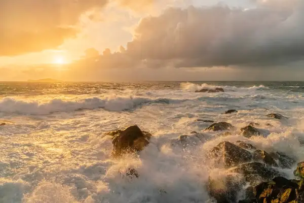 Sunset over a turbulent ocean with waves crashing against rocky formations. The sky is filled with dramatic clouds, illuminated in shades of orange and gold by the setting sun. A distant landmass is silhouetted on the horizon.