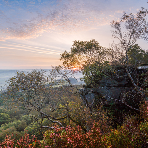 Edge of autum: A sunrise illuminates a countryside vista from a high vantage point. Various hues of pinks and blues adorn the sky. In the foreground are rocky outcrops and silhouettes of twisted trees. Below, layers of greenery blanket rolling hills, hinting at a tranquil rural landscape.