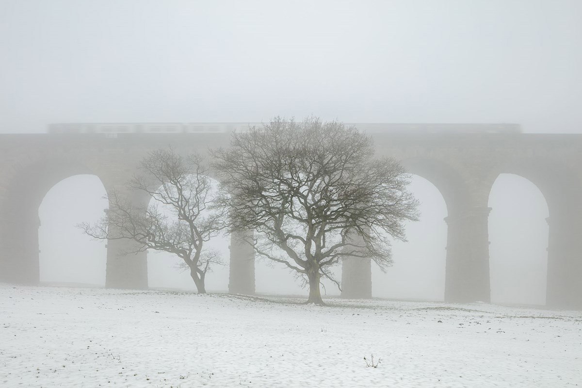 A train passes over the Crimple Valley Viaduct on a cold winter day  a building covered in snow