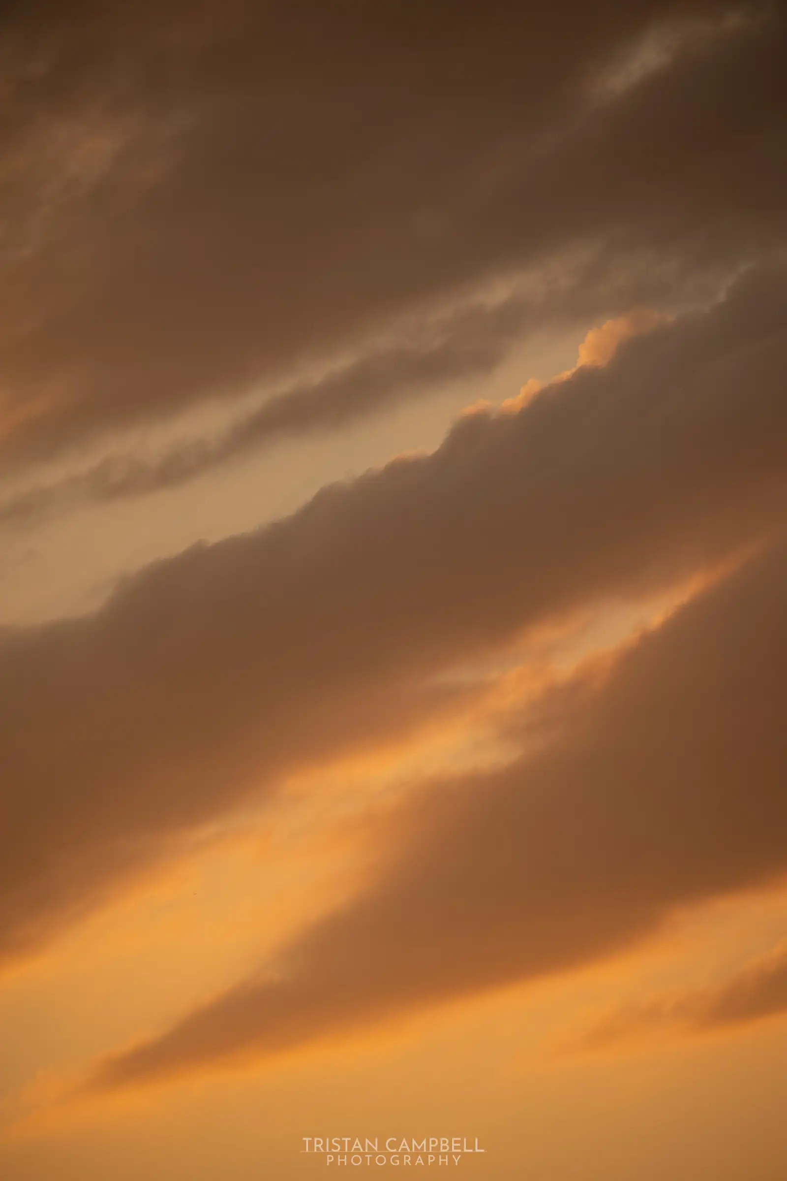 Golden sunset sky with thick, elongated clouds. The clouds are tinged with shades of warm orange and brown, creating a serene, atmospheric scene.