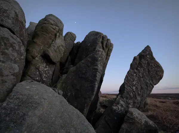 Large, weathered rock formations standing against a clear evening sky with a small crescent moon visible. The rugged rocks have a textured surface, and the landscape beyond is a flat expanse of grassland, tinged with a hint of pink from the setting sun.
