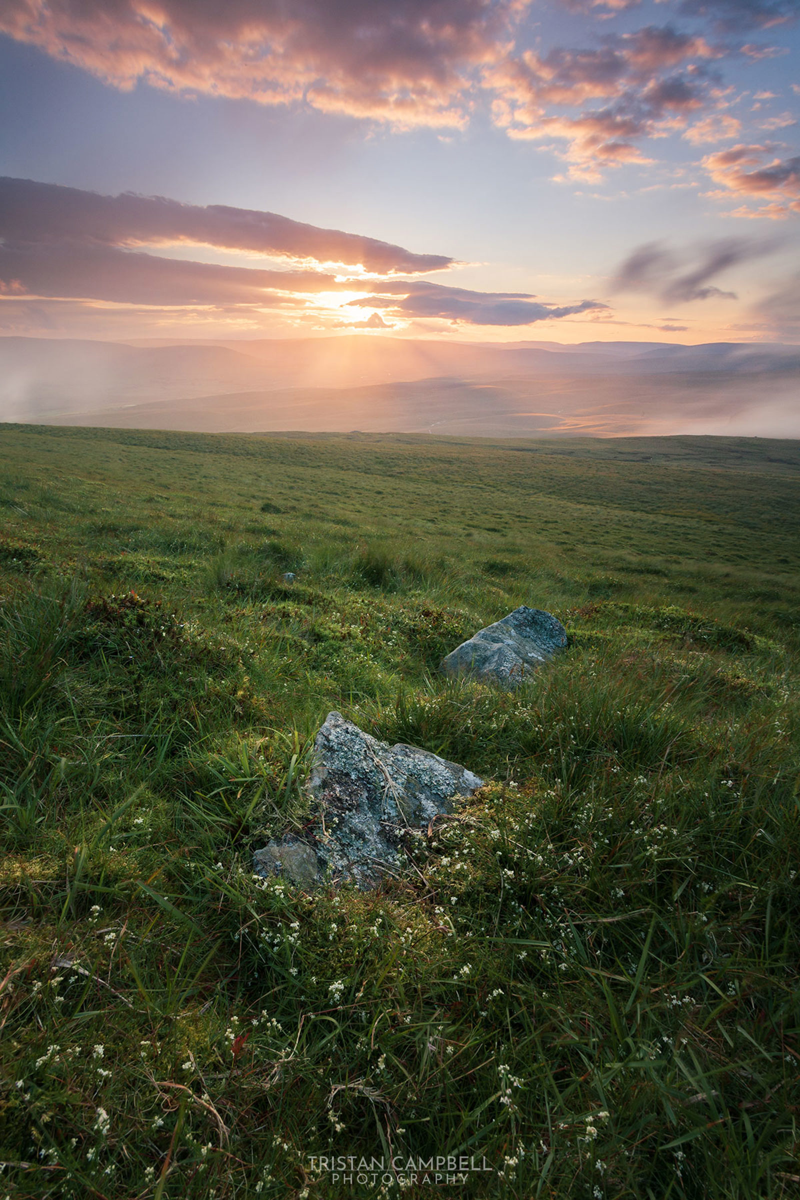 Sunset over a vast green landscape with rolling hills. Sun rays break through scattered clouds, casting a warm glow across the scene. In the foreground, a few large rocks sit amidst grass and small white flowers, adding texture to the area.