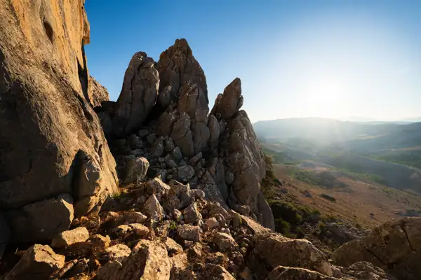 Rugged rocky outcrops sharply rise in the foreground, casting shadows in the warm light of the setting sun. The landscape extends into a vast valley with rolling hills and scattered greenery under a clear blue sky. Sunlight streams across the scene, creating a serene and expansive view.