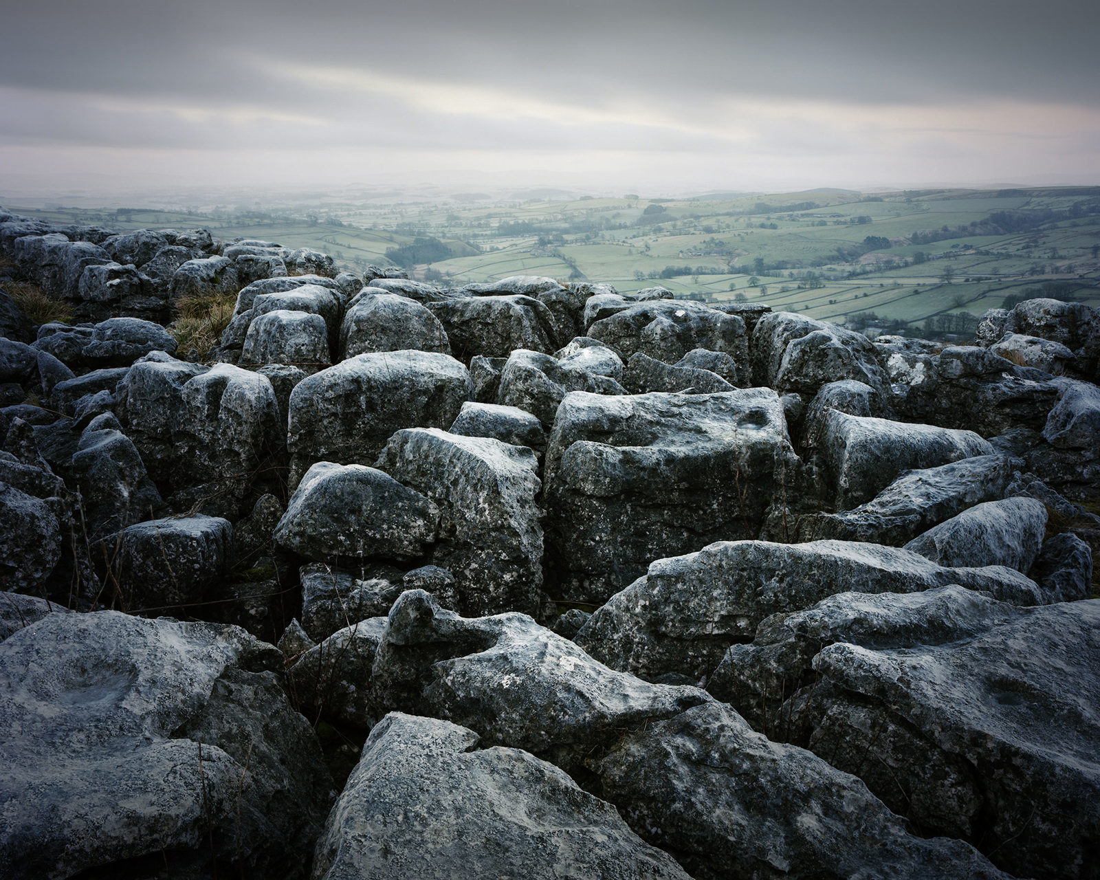 A rugged limestone pavement dominates the foreground, with large, weathered rocks displaying distinctive clints and grikes. In the background, a lush, rolling landscape stretches under an overcast sky, with muted patches of green fields and distant hills fading into the horizon.