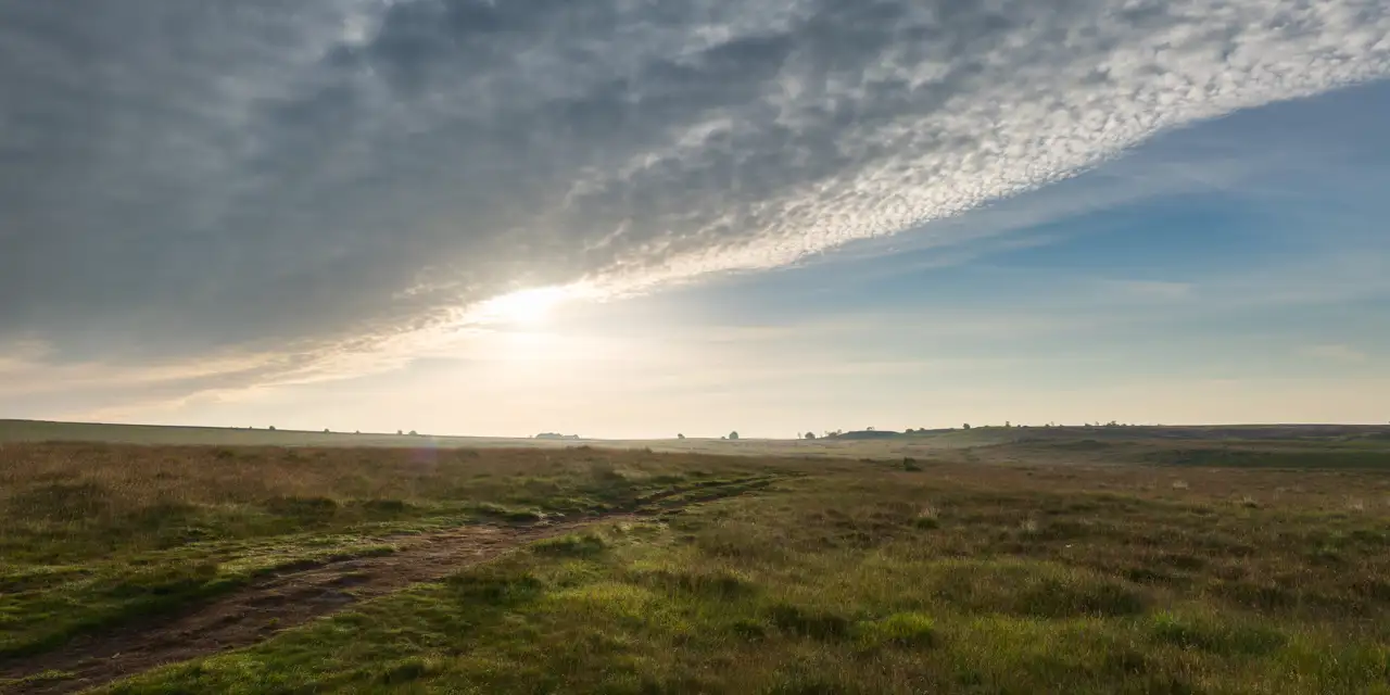 Wide open grassland under a partly cloudy sky with the sun low on the horizon, casting a warm glow across the landscape. A dirt path runs through the grass, leading towards distant, low hills. The sky transitions from grey clouds on the left to a clearer blue on the right.