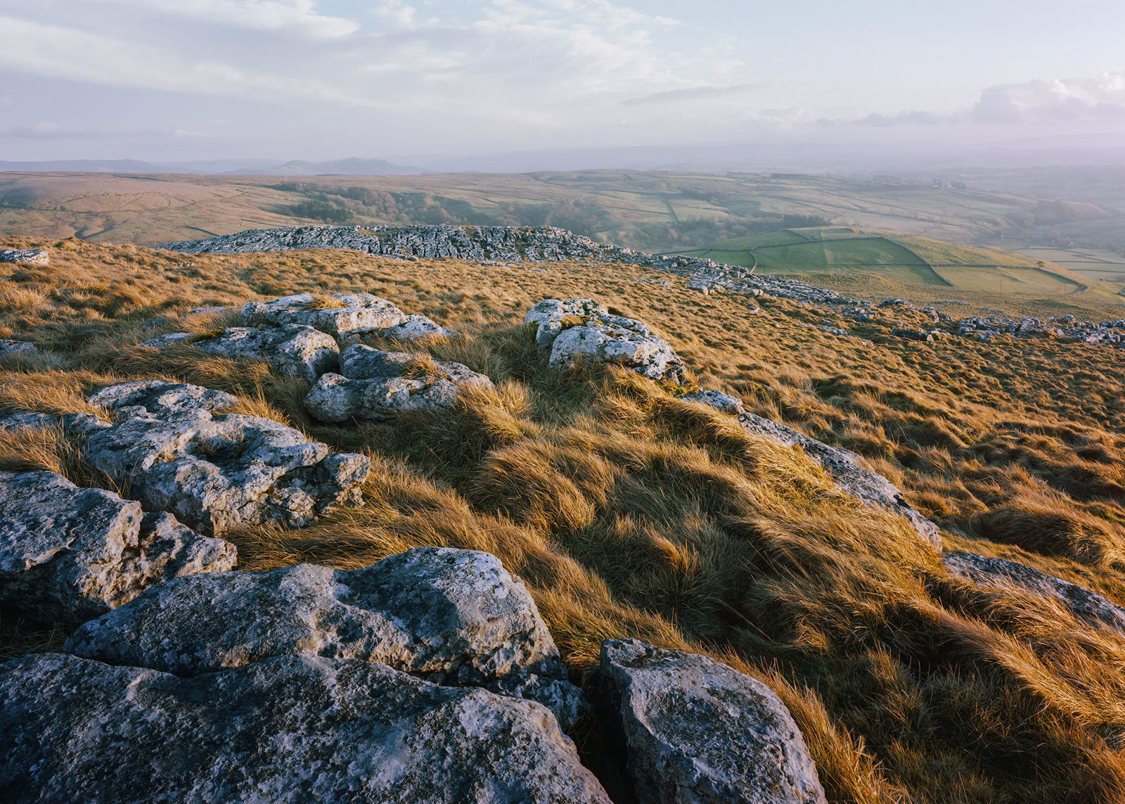Rugged landscape with scattered grey rocks and uneven grassy terrain bathed in soft sunlight. The background reveals a vast, patchwork of fields and rolling hills under a partly cloudy sky.