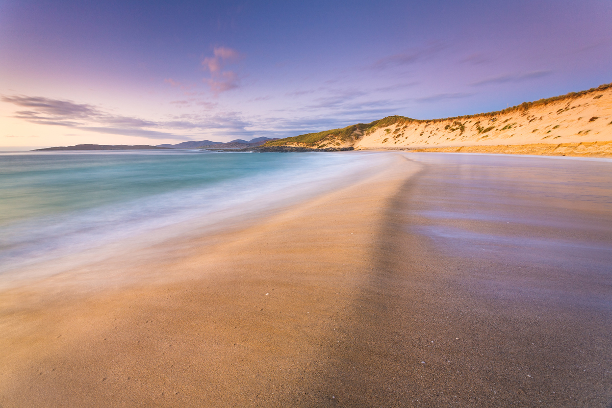 A sweeping sandy beach graces the foreground, with gentle waves caressing the shore and leaving a smooth, wet surface. A grass-topped dune rises to the right, bathed in warm light from a softly lit sky streaked with lavender and gold hues. In the distance, gentle hills fade into the horizon under a tranquil sky. a beach with sand and water