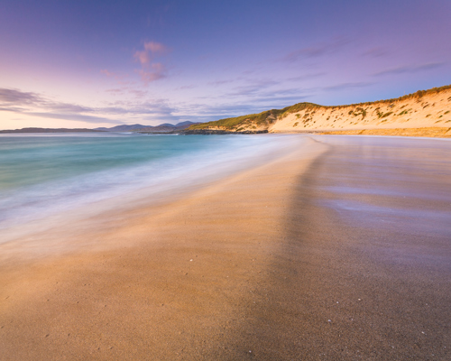 Seascapes of the Outer Hebrides: A sweeping sandy beach graces the foreground, with gentle waves caressing the shore and leaving a smooth, wet surface. A grass-topped dune rises to the right, bathed in warm light from a softly lit sky streaked with lavender and gold hues. In the distance, gentle hills fade into the horizon under a tranquil sky. a beach with sand and water