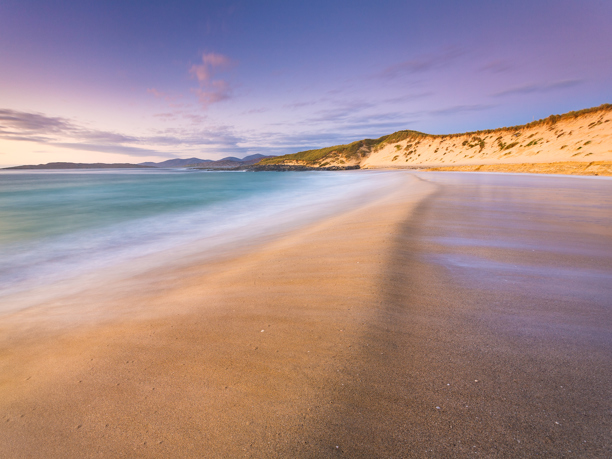 Seascapes of the Outer Hebrides A sweeping sandy beach graces the foreground, with gentle waves caressing the shore and leaving a smooth, wet surface. A grass-topped dune rises to the right, bathed in warm light from a softly lit sky streaked with lavender and gold hues. In the distance, gentle hills fade into the horizon under a tranquil sky.