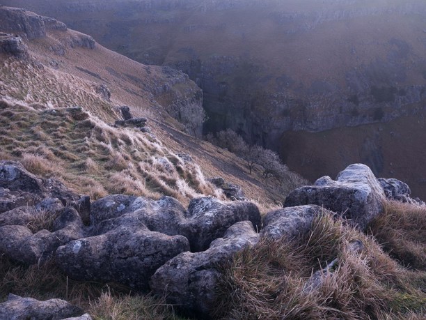 Yorkshire Dales: A rugged landscape with windswept grasses and lichen-covered rocks in the foreground. Beyond lies a deep limestone ravine edged by sheer cliffs, under a dusky sky in Yorkshire Dales.