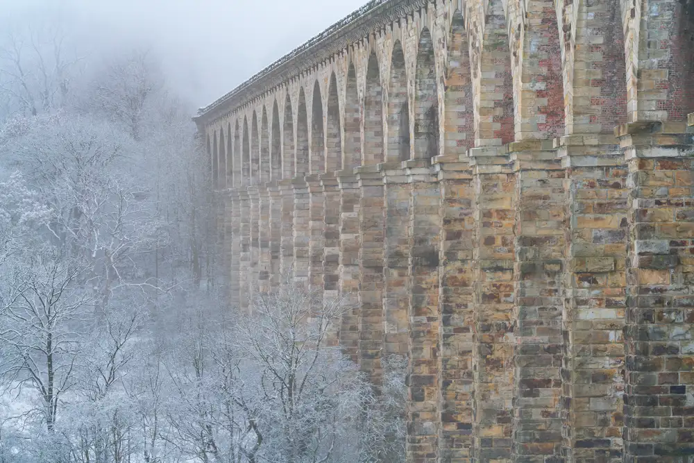 Tall stone viaduct with numerous archways extends into a misty, wintry scene. Snow-covered trees surround the structure, and light snow blankets the ground, creating an ethereal atmosphere.