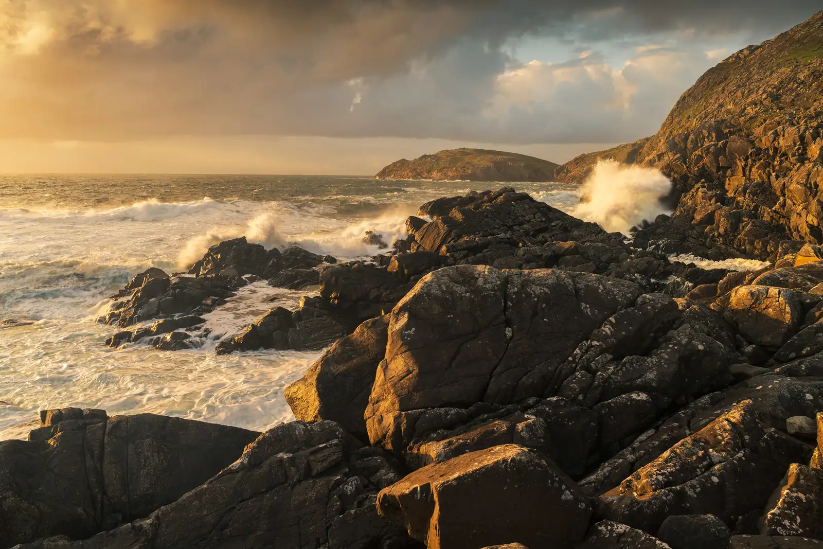Rocky coastal landscape with waves crashing against dark, jagged rocks. The scene is bathed in warm, golden light from a setting or rising sun, casting long shadows. In the background, an ocean stretches to the horizon under a partly cloudy sky, with a distant landform visible.