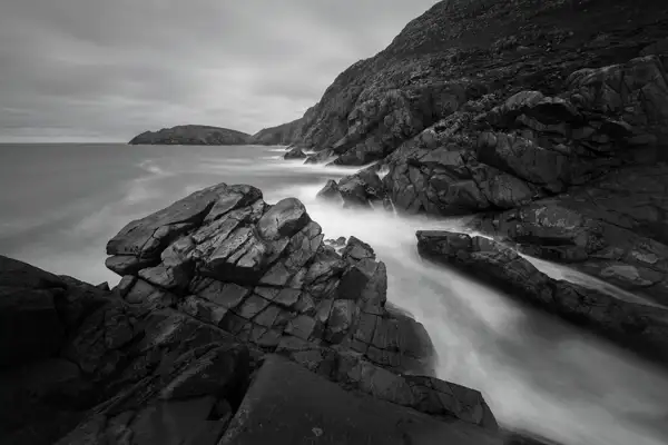 Rocky coastline with dramatic cliffs and large boulders jutting into the sea. The water appears smooth and misty, suggesting a long exposure shot. Overcast sky creates a moody atmosphere, with the distant coastline visible in the background. The image is in black and white, highlighting textures and contrasts.