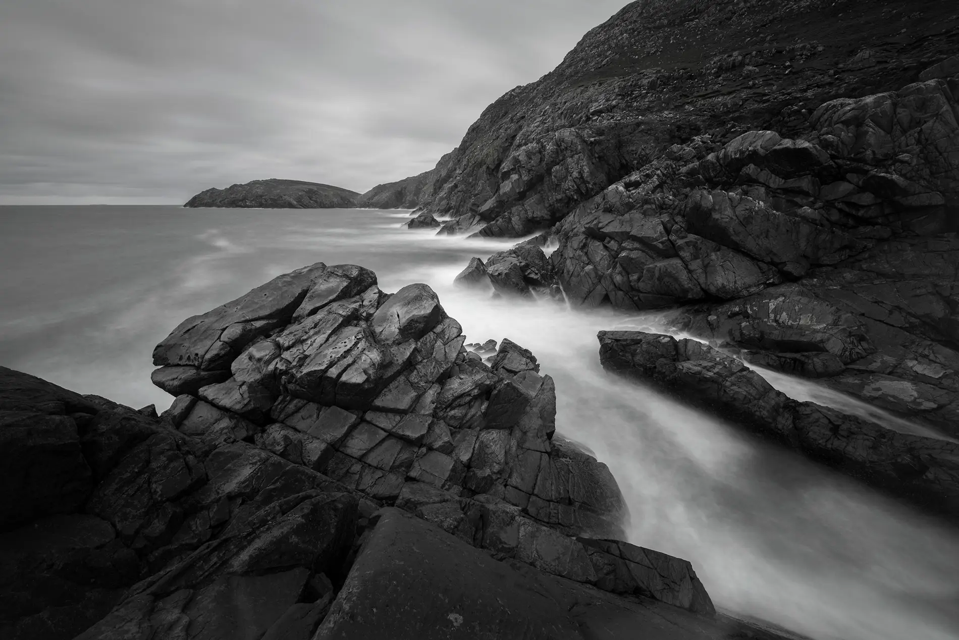 Jagged coastal rocks with mist-soft surf flowing into a sheltered cove beneath an overcast sky, in black and white