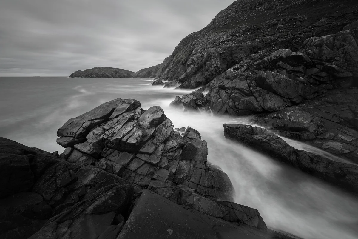Jagged coastal rocks with mist-soft surf flowing into a sheltered cove beneath an overcast sky, in black and white