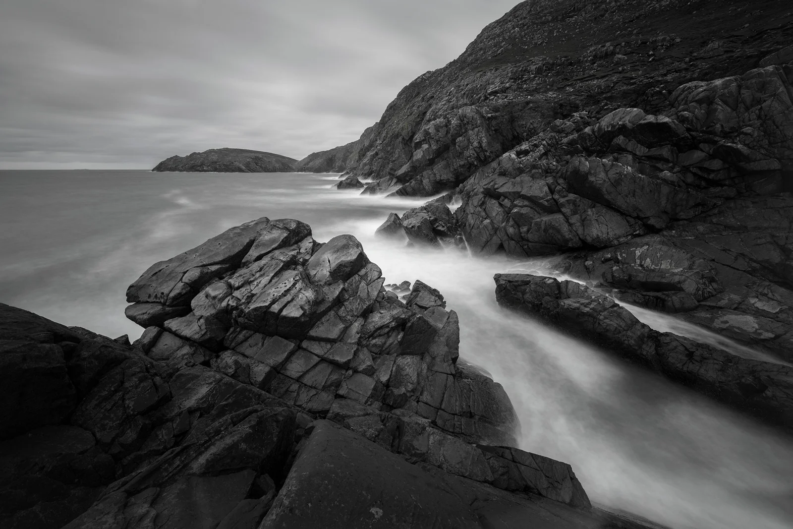 Jagged coastal rocks with mist-soft surf flowing into a sheltered cove beneath an overcast sky, in black and white
