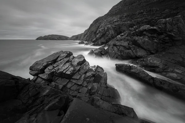 Jagged coastal rocks with mist-soft surf flowing into a sheltered cove beneath an overcast sky, in black and white