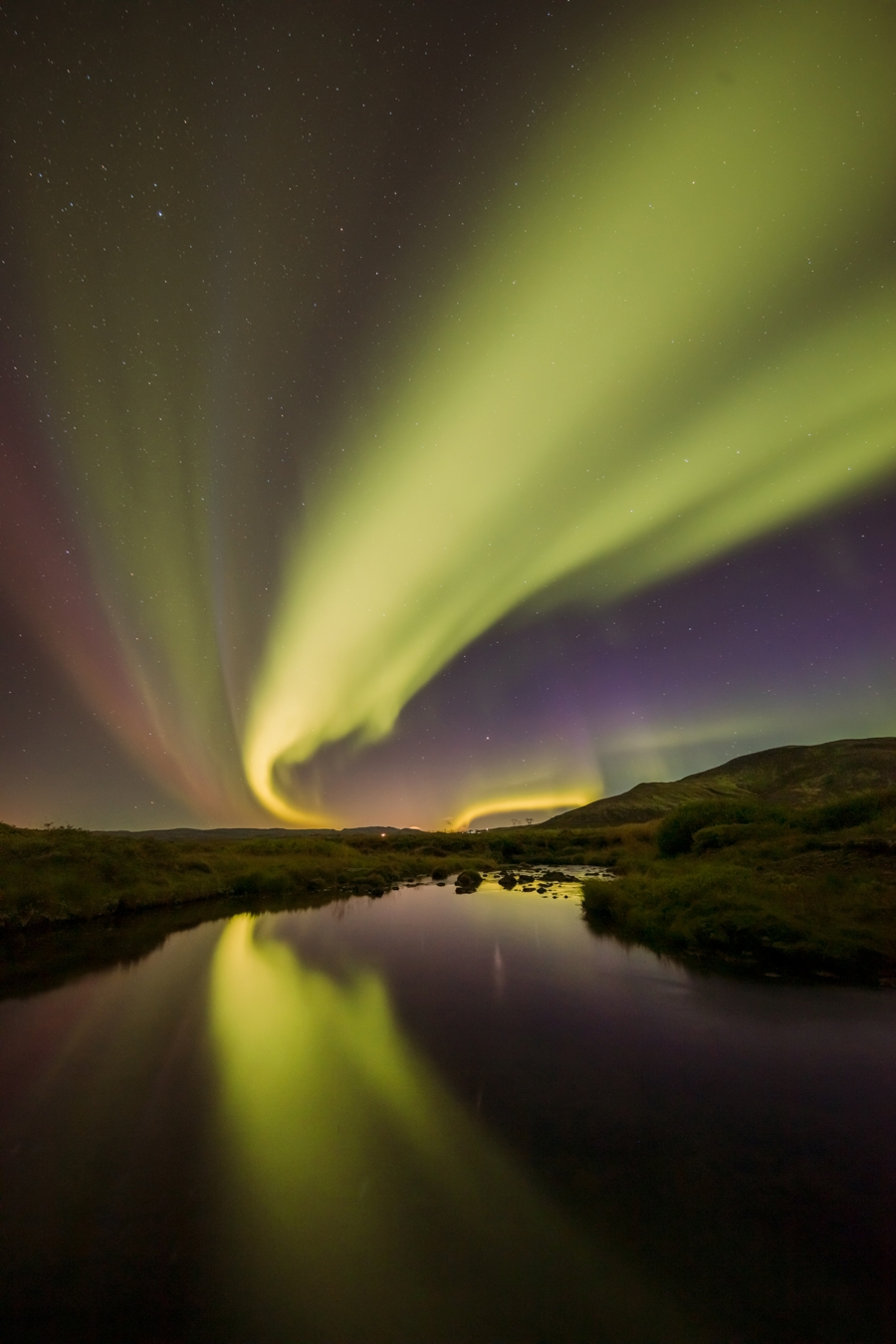 Northern Lights in Iceland displaying a mesmerizing array of greens and purples that arc across a star-filled night sky, with their reflection shimmering on the surface of a tranquil river below. Gentle hills frame the scene. a sky view looking up at the camera