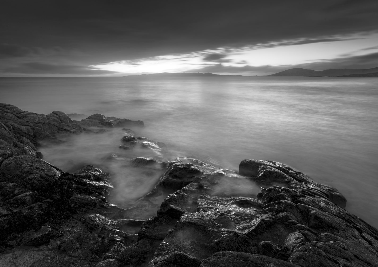 A monochrome seascape captures a serene atmosphere. Jagged rocks adorn the foreground, as soft mist-like water laps around them, likely from a slow shutter speed. The horizon is subtle under a gradient sky, hinting at a setting or rising sun peeking through clouds. Faraway mountains complete the tranquil scene.