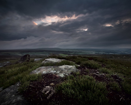 Moorland Landscapes: Moody North Yorkshire landscape at dusk. Foreground shows rugged terrain with rocks and tufts of grass. Dark, brooding clouds dominate the sky with a sliver of warm light piercing through. Distant hills fade into the horizon. a person standing on a grassy hill