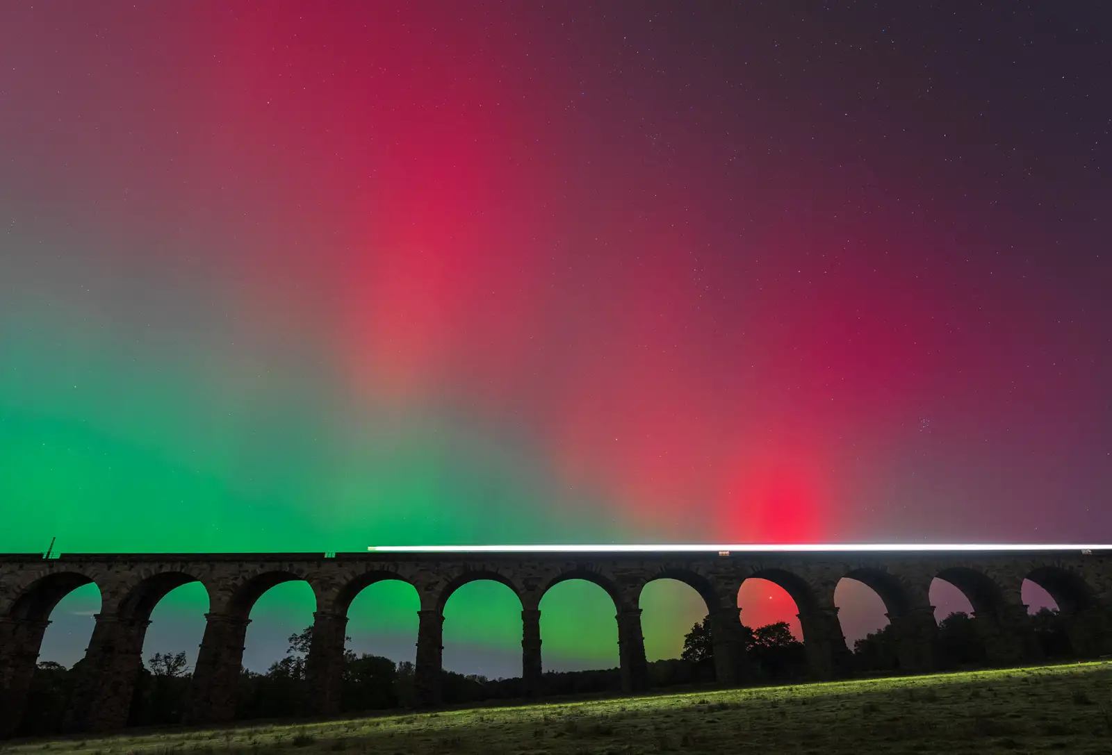 A stone viaduct with multiple arches spans across the foreground, silhouetted against the vibrant aurora borealis in the night sky. Bands of red and green light create a dramatic effect above, while a faint trail of light suggests a passing train along the top of the viaduct. Stars are visible in the sky, and the landscape below is dimly lit.