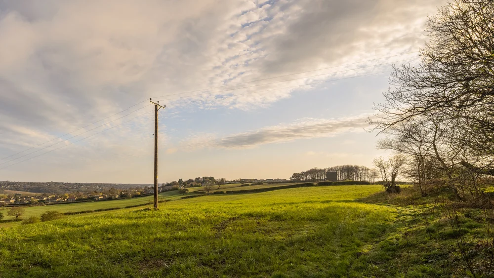 Wooden telegraph pole in a green hillside field with a village and trees below at golden hour in early spring