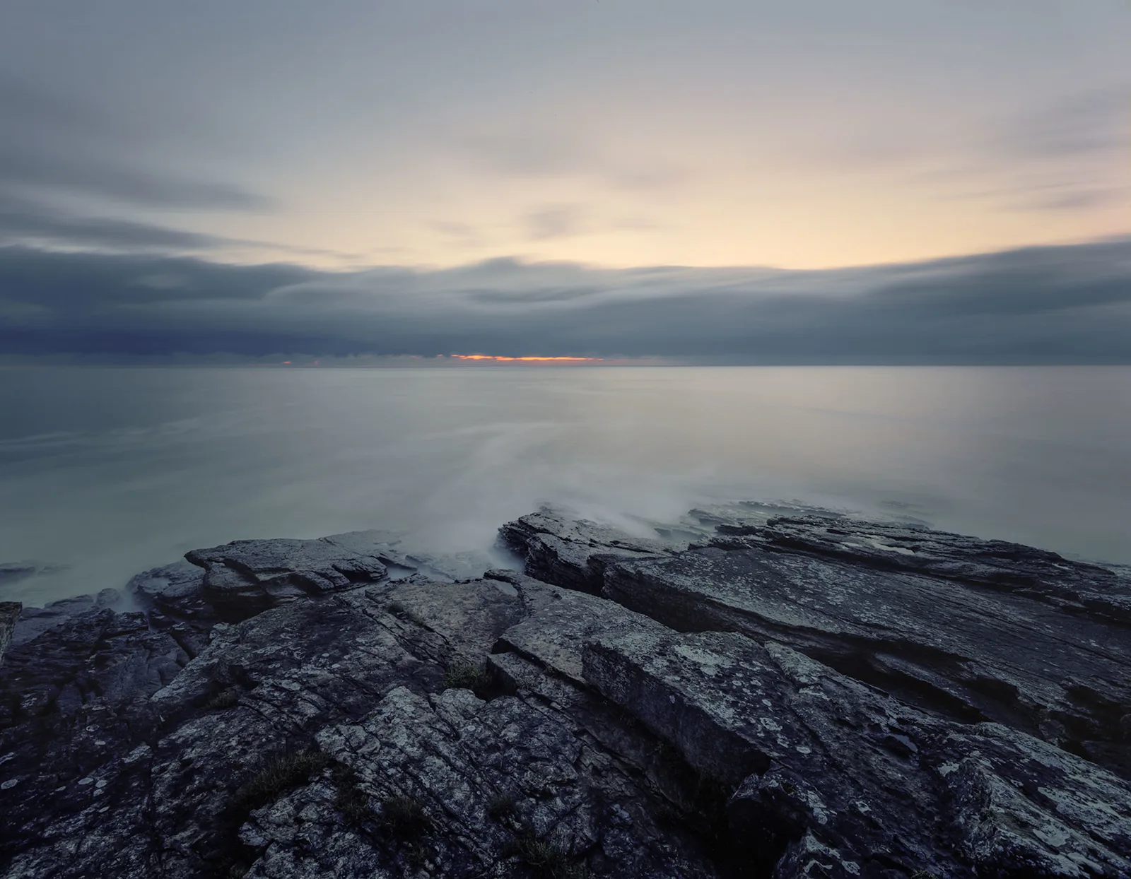 Layered dark rocks in foreground with calm sea and distant mountains beneath overcast sky at dusk.
