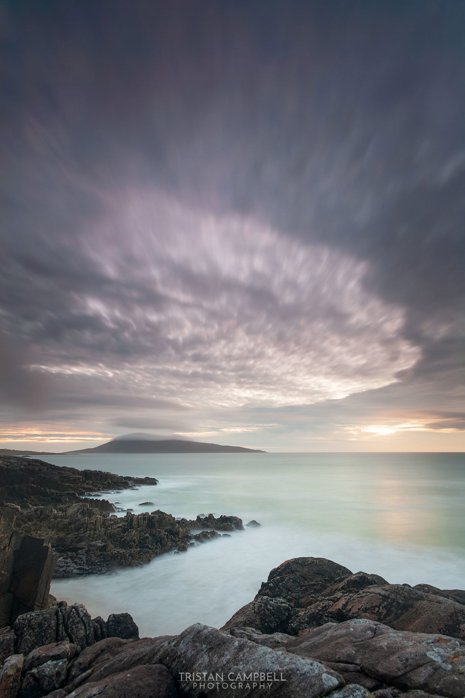 Rocky coastline with blurred waves crashing against dark, rugged rocks. In the distance, a misty mountain is partially obscured by clouds. The sky is dramatic with swirling, soft grey and purple clouds, illuminated by a subtle, warm sunset glow on the horizon.