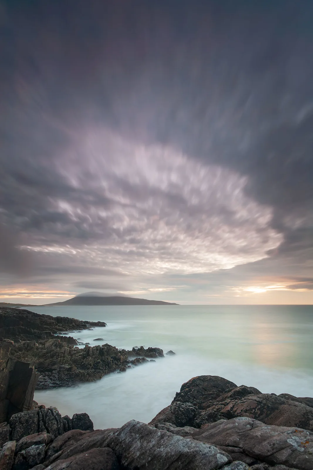 Rocky coastline with misty sea and distant volcanic island under dramatic cloudy sky at dusk.