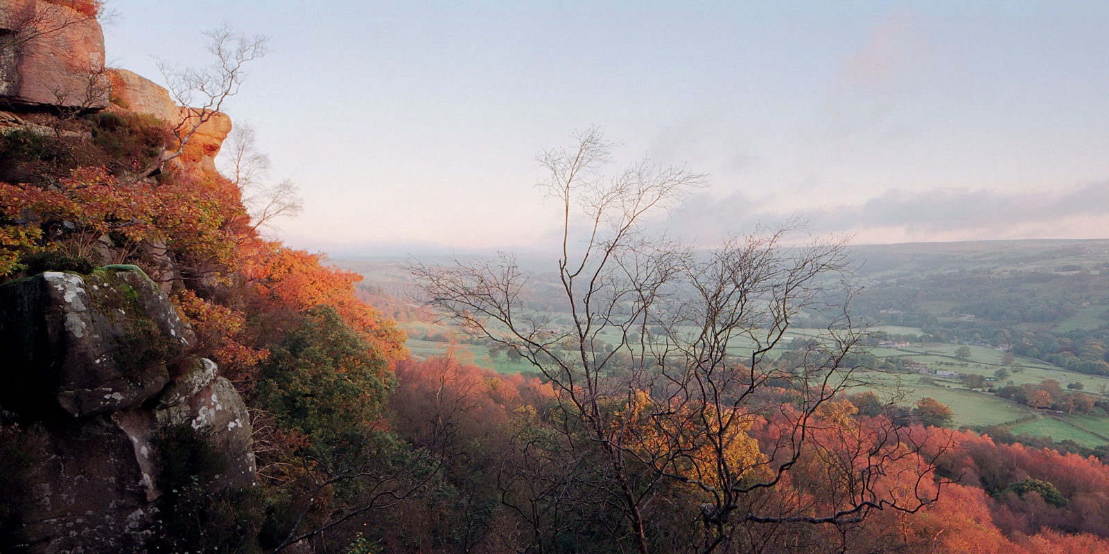 A rocky cliff adorned with autumnal foliage overlooks a vast, rolling valley. The trees display vibrant hues of orange and red, while the valley below stretches into the distance with green fields and scattered trees. The sky above is pale with soft clouds, giving a serene atmosphere to the landscape.