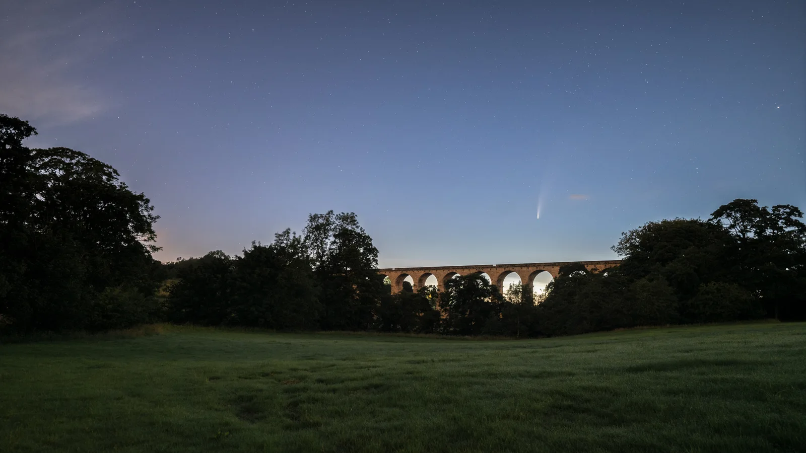 Stone viaduct with multiple arches spanning wooded valley at dusk, starlit sky above, grass field in foreground.